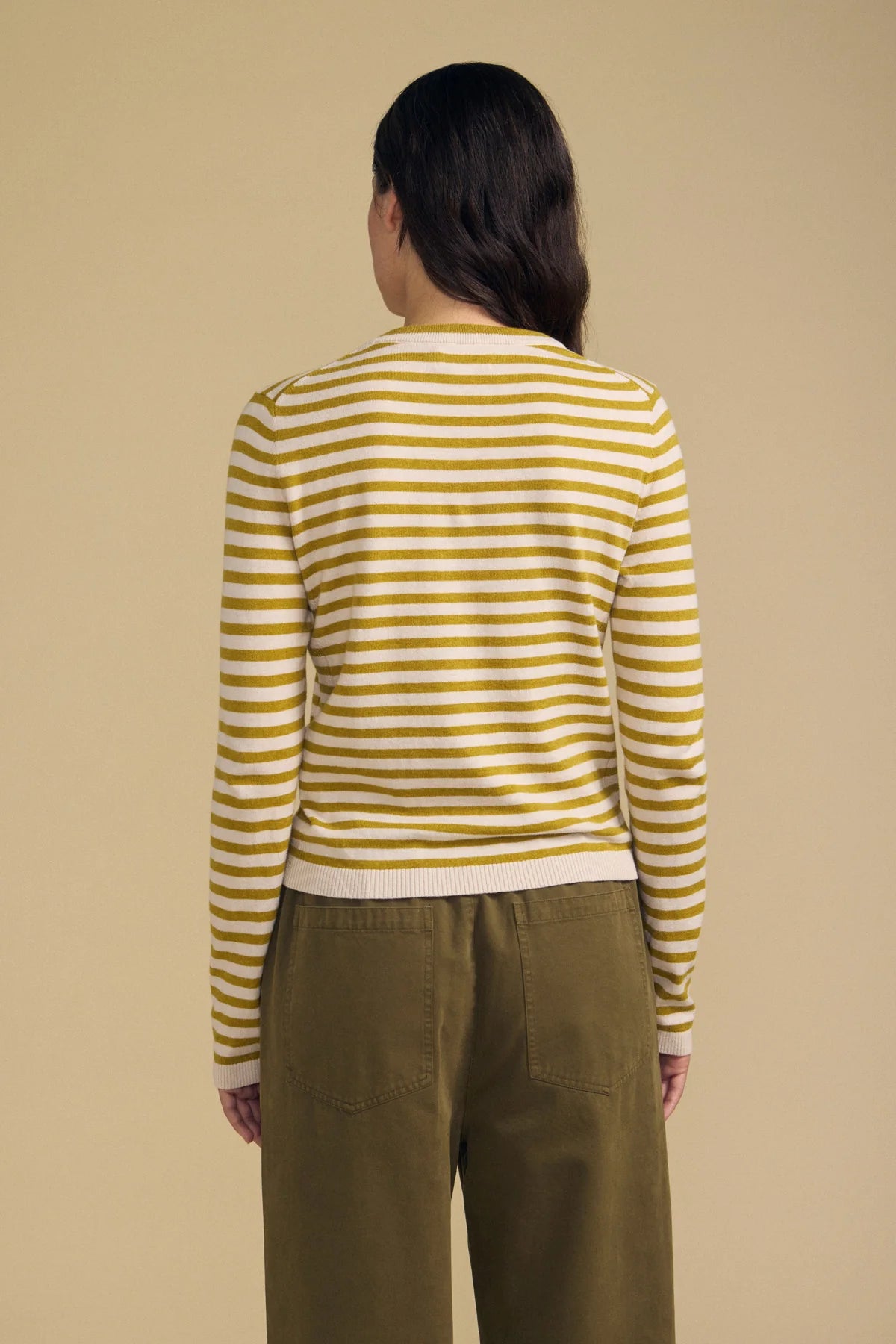 A model shown from the back wearing a yellow and white striped long-sleeve knit top and olive green pants, set against a solid beige background.