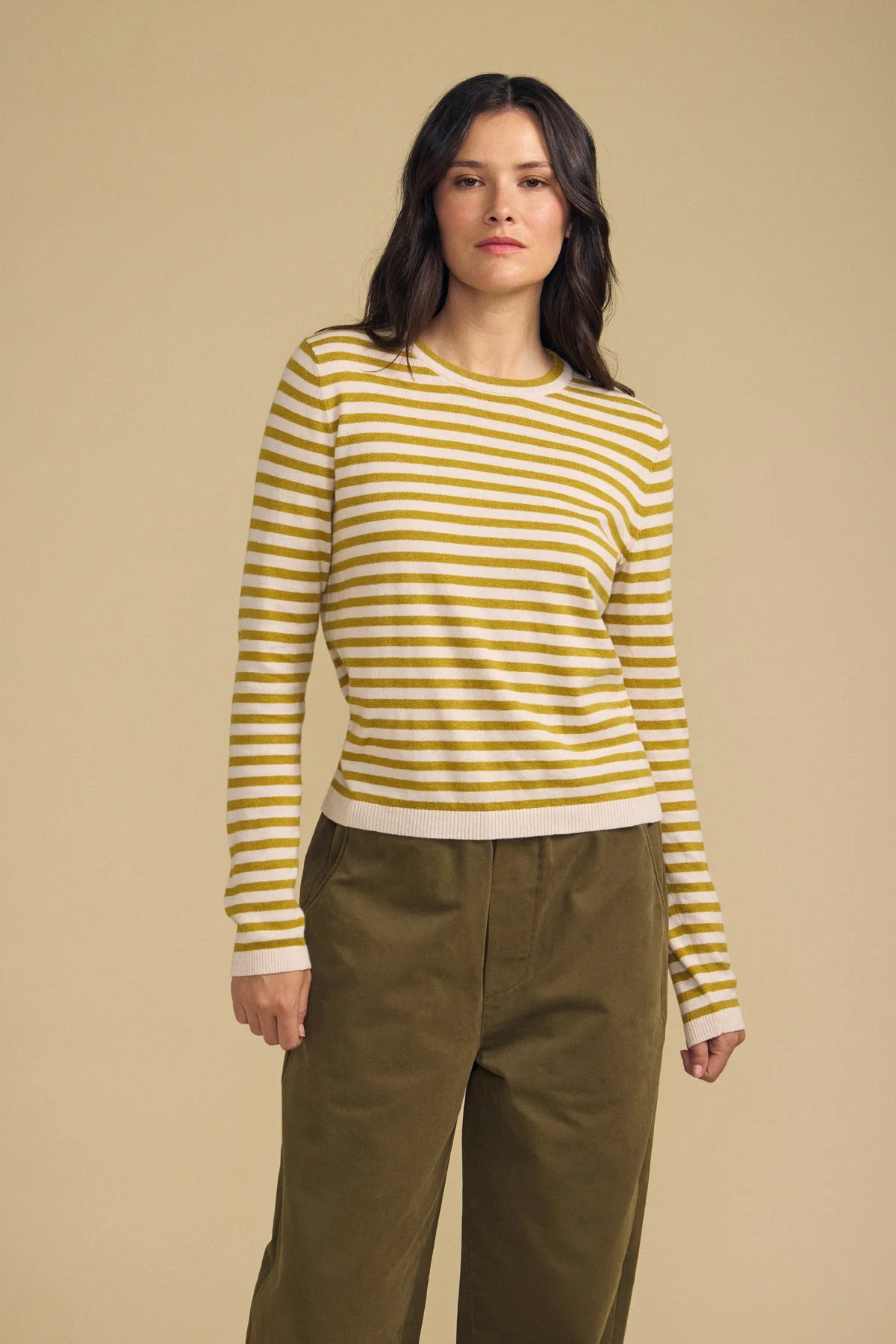 A woman modeling a yellow and white striped long-sleeve knit top with olive green pants, standing against a solid beige studio background.