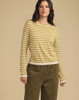 A woman modeling a yellow and white striped long-sleeve knit top with olive green pants, standing against a solid beige studio background.