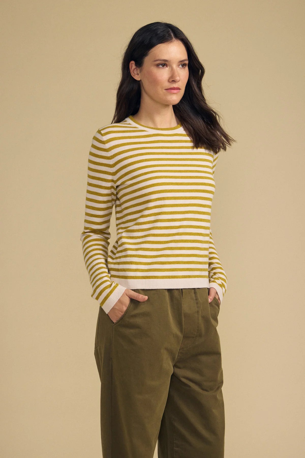 A woman modeling a yellow and white horizontally striped long-sleeve knit top with olive green pants, standing against a solid beige studio background.