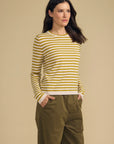 A woman modeling a yellow and white horizontally striped long-sleeve knit top with olive green pants, standing against a solid beige studio background.