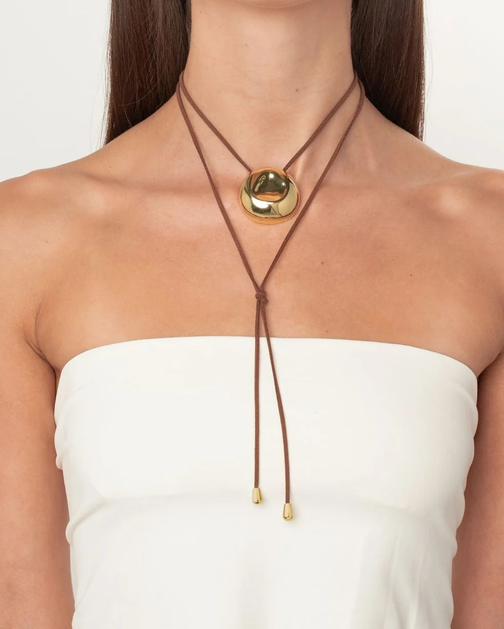 A close-up of a model wearing a brown double-strand cord necklace with a large, polished gold oval pendant and small gold bead accents at the ends, styled with a white strapless top.
