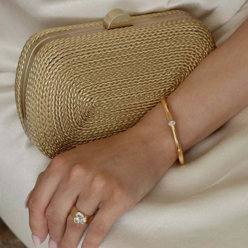 Gold clutch held by a hand with a gold ring and bracelet on a neutral background