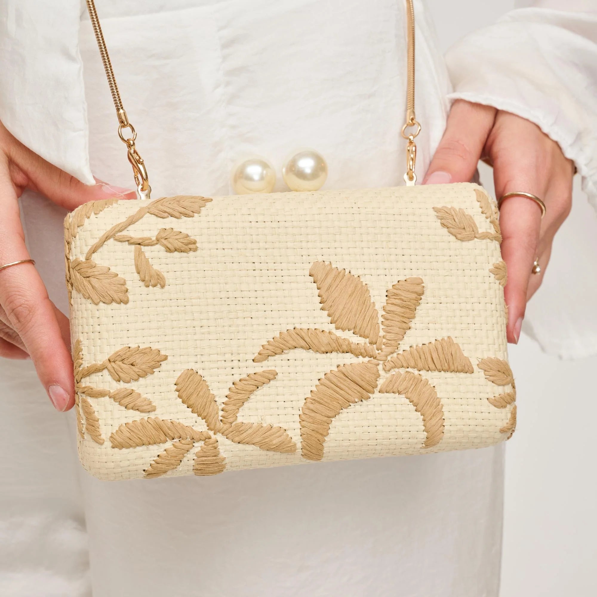 Beige handbag with floral embroidery held by a person wearing a white outfit.