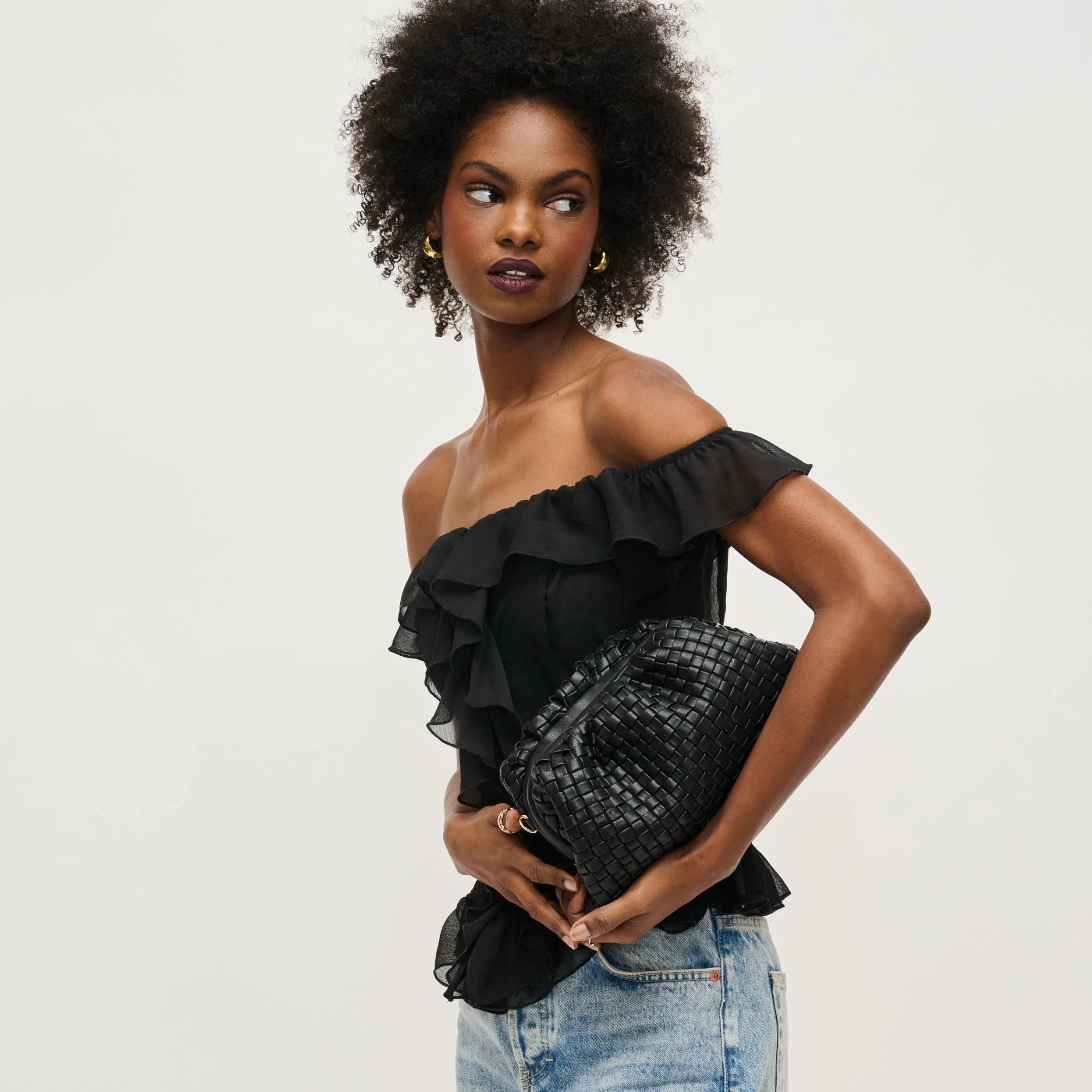 Woman wearing a black off-shoulder top and denim shorts, holding a black textured bag on a light gray background.