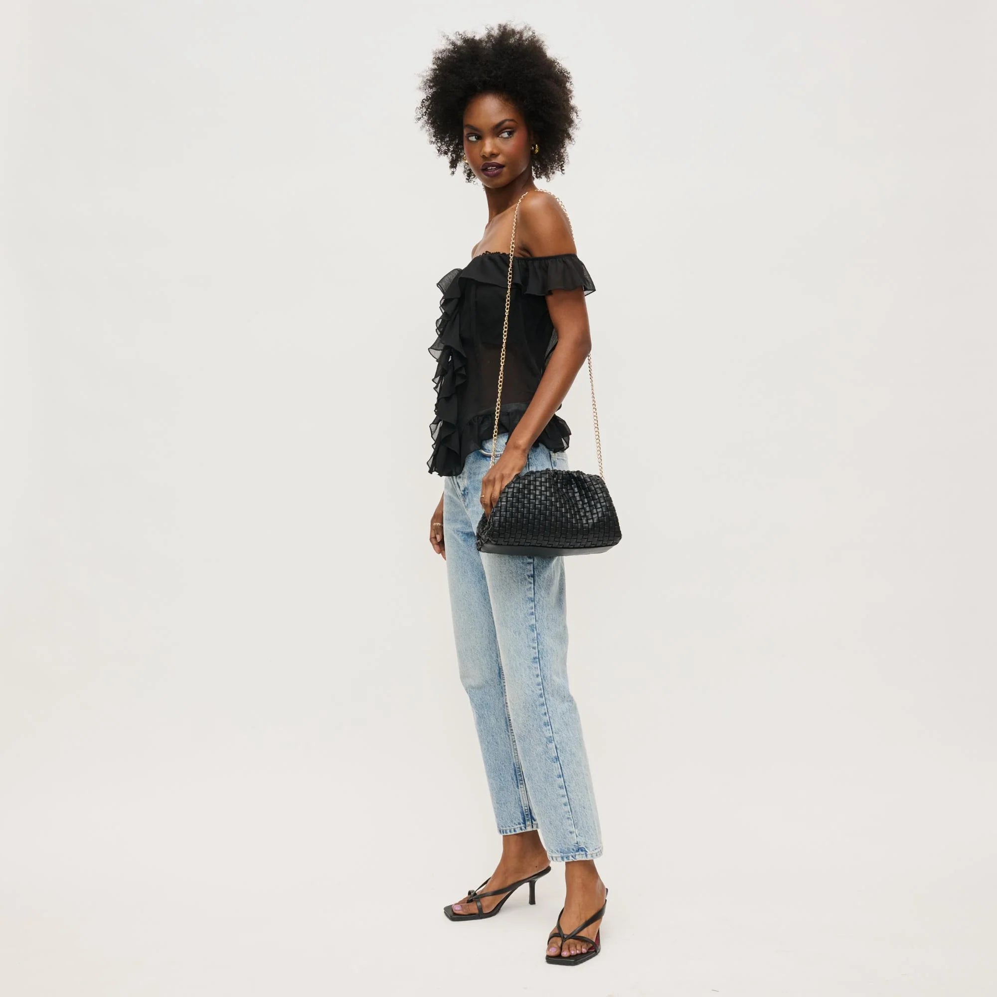 Woman wearing a black off-shoulder top and light blue jeans, holding a black handbag on a white background