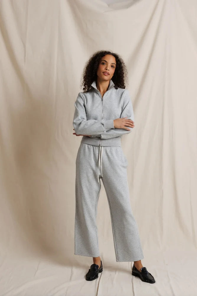A woman modeling a light gray heathered two-piece loungewear set consisting of a quarter-zip long-sleeve top and matching wide-leg sweatpants, standing against a draped beige fabric backdrop.