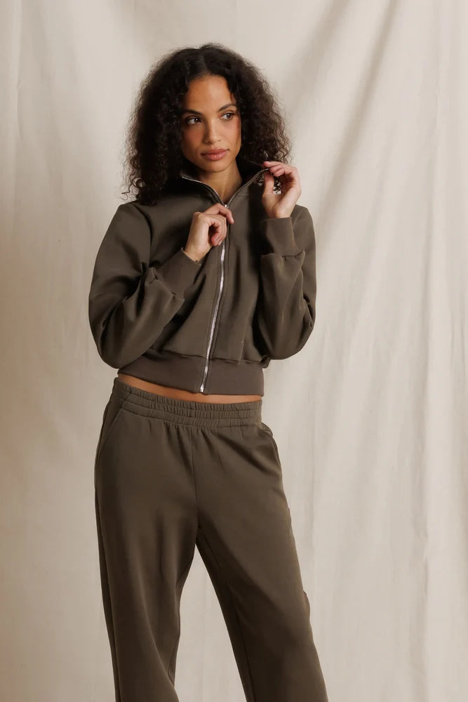 A woman modeling a matching dark brown cropped zip-up hoodie and sweatpants set against a draped beige fabric backdrop.