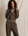 A woman modeling a matching dark brown cropped zip-up hoodie and sweatpants set against a draped beige fabric backdrop.