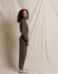 A woman with curly dark hair stands in profile wearing a matching taupe-brown long-sleeve top and slim trousers, paired with white loafers, against a draped off-white fabric backdrop.
