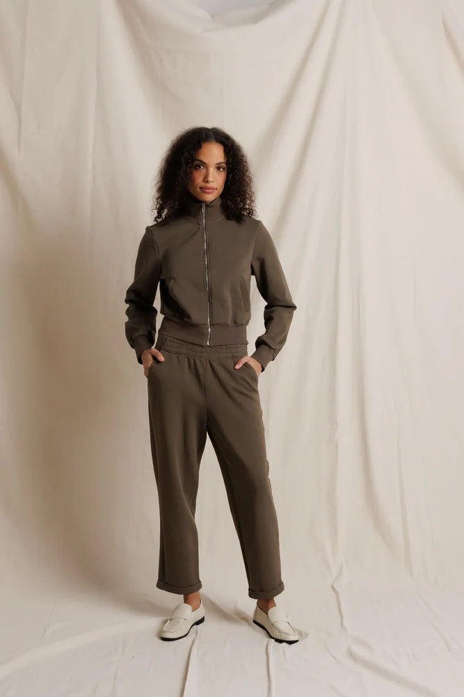A woman modeling a matching two-piece tracksuit in a muted olive-brown color, consisting of a zip-up long-sleeve top and matching pants, standing against a draped off-white fabric backdrop.