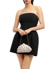 A woman wearing a strapless black mini dress and holding a white clamshell-shaped handbag with a gold chain strap, photographed against a plain white background.