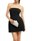 A woman wearing a strapless black mini dress and holding a white clamshell clutch, photographed against a plain white background.