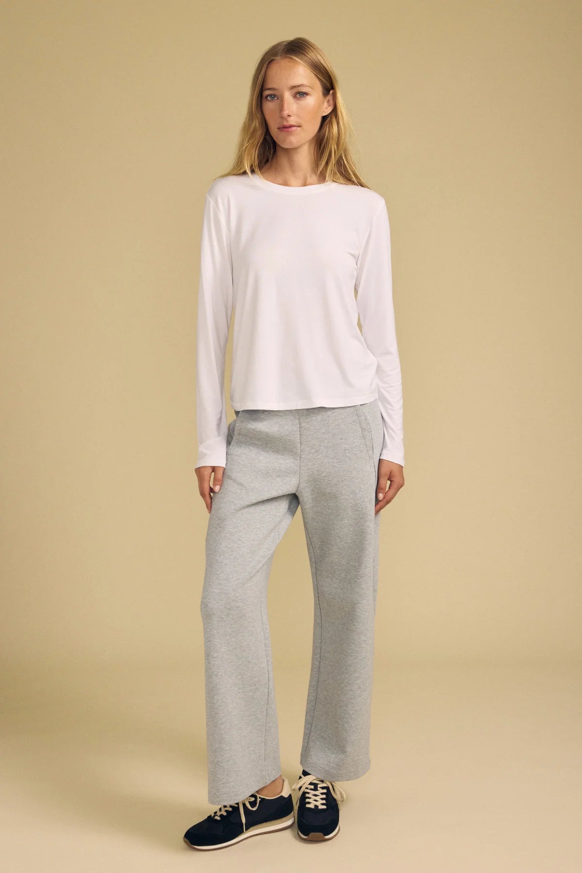 A woman modeling a white long-sleeve crewneck top and light gray wide-leg sweatpants, paired with navy blue sneakers, against a solid beige studio background.