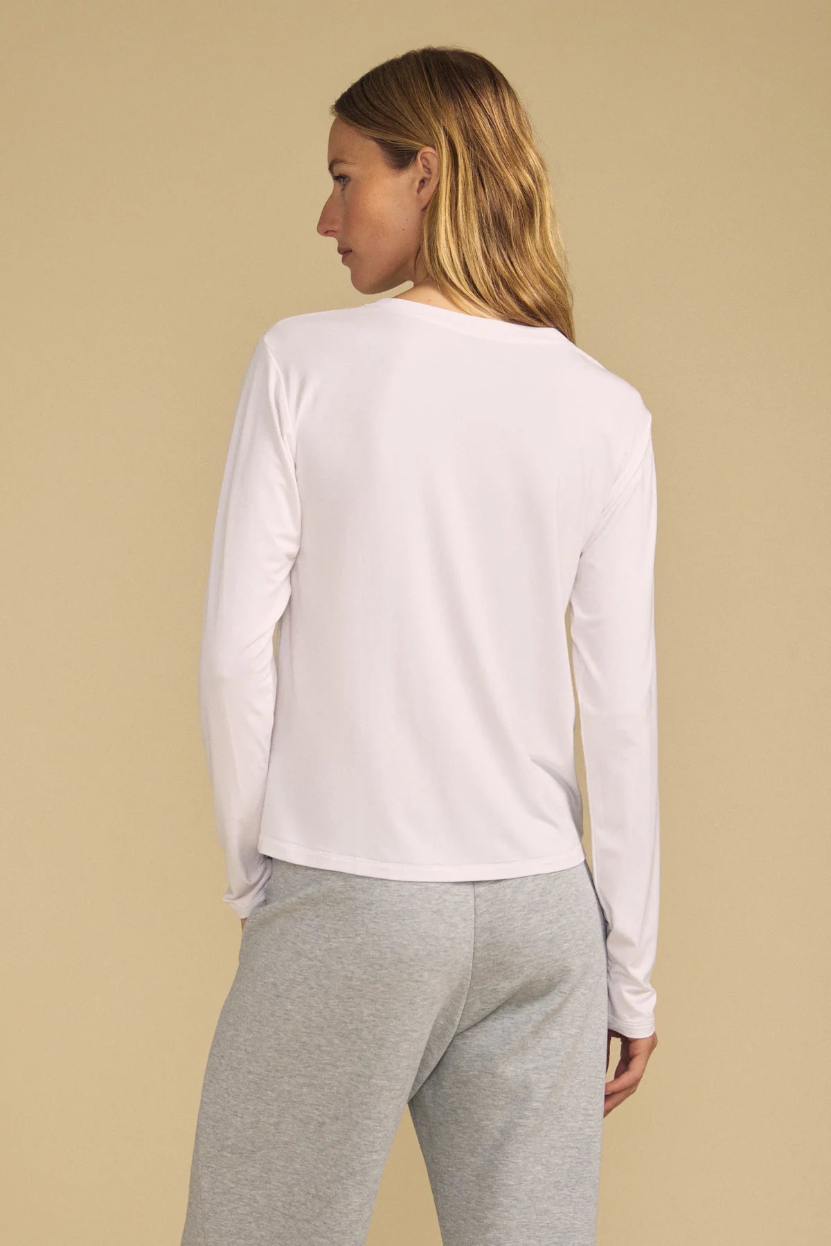 A woman shown from the back wearing a white long-sleeve top and light gray sweatpants, standing against a solid beige studio background.