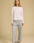 A woman modeling a white long-sleeve crewneck top and light gray wide-leg sweatpants, paired with navy blue sneakers, against a solid beige studio background.