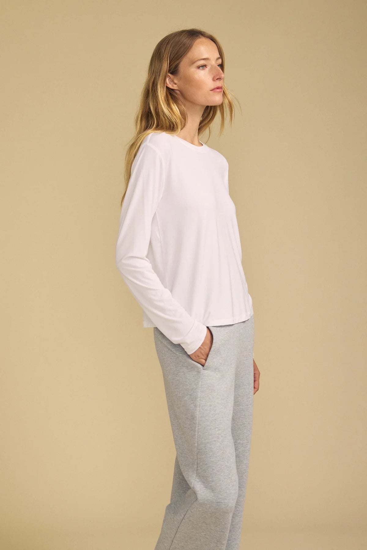 A woman in a white long-sleeve top and light gray sweatpants, shown in profile against a solid beige studio background.