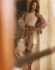 Woman standing in a softly lit room wearing a white top and beige pants.