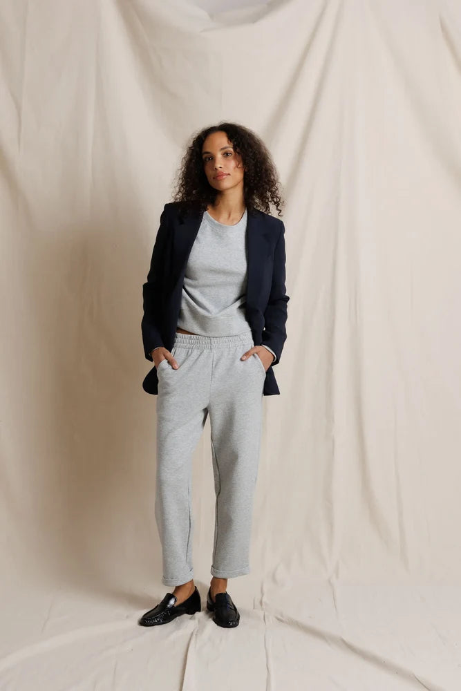 Woman wearing a dark blazer, light gray top, and pants against a plain background