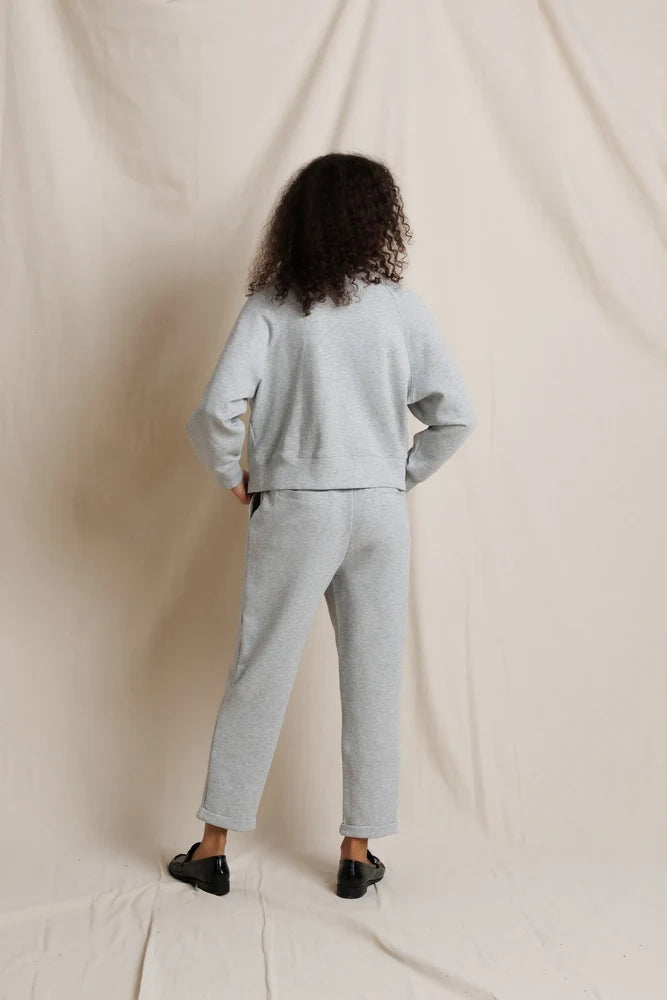 A model viewed from behind wearing a light gray heather sweatshirt and matching sweatpants, standing against a draped cream fabric backdrop.