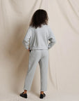 A model viewed from behind wearing a light gray heather sweatshirt and matching sweatpants, standing against a draped cream fabric backdrop.