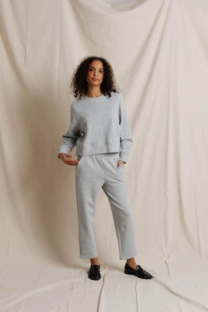 A woman modeling a light gray two-piece loungewear set consisting of a cropped long-sleeve sweatshirt and matching straight-leg sweatpants, paired with black loafers, set against a draped cream fabric backdrop.