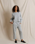 A woman modeling a light gray two-piece loungewear set consisting of a cropped long-sleeve sweatshirt and matching straight-leg sweatpants, paired with black loafers, set against a draped cream fabric backdrop.