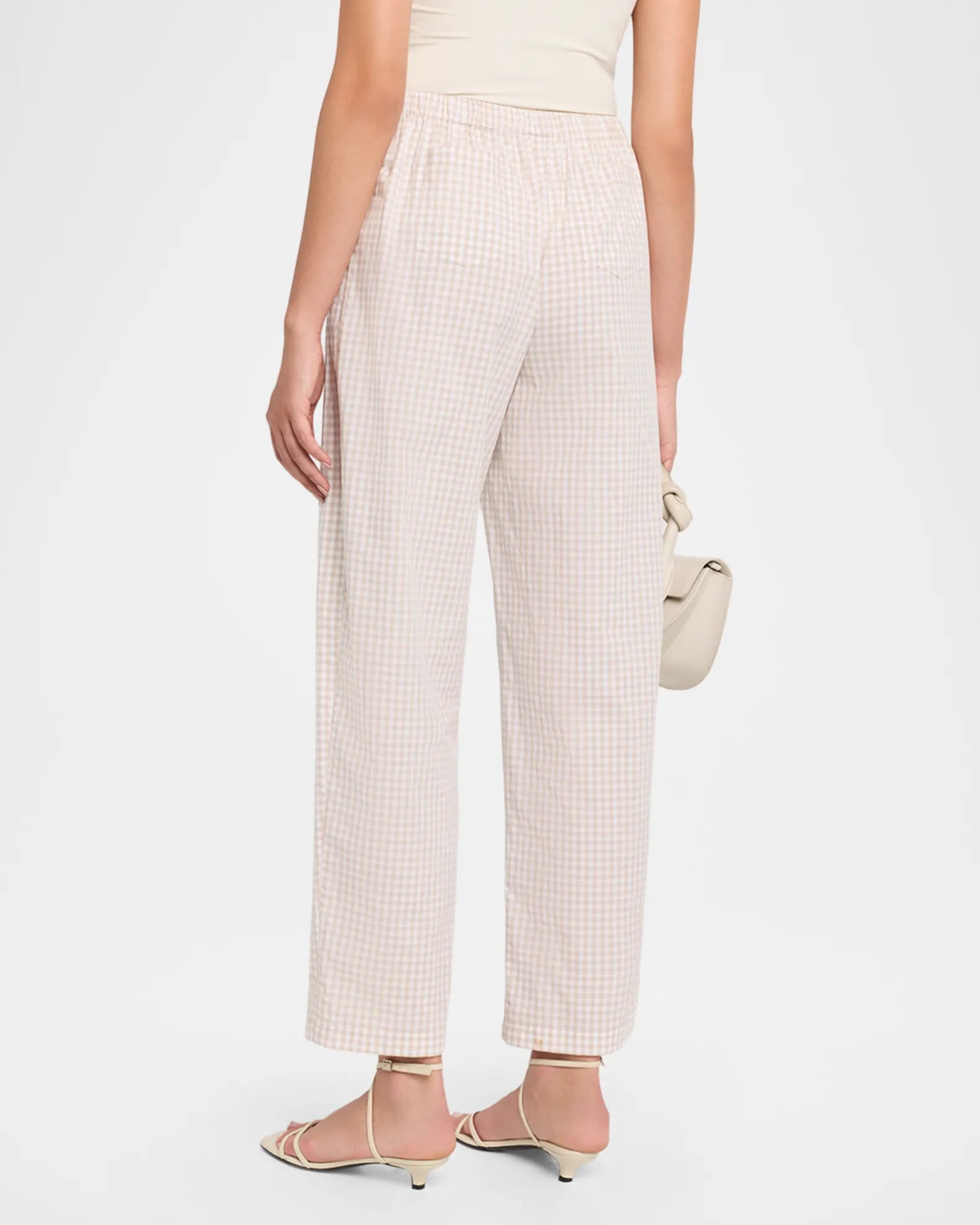 A model shown from the back wearing light beige wide-leg cropped trousers with a small white gingham pattern, paired with beige strappy sandals and holding a matching beige handbag against a plain white studio background.