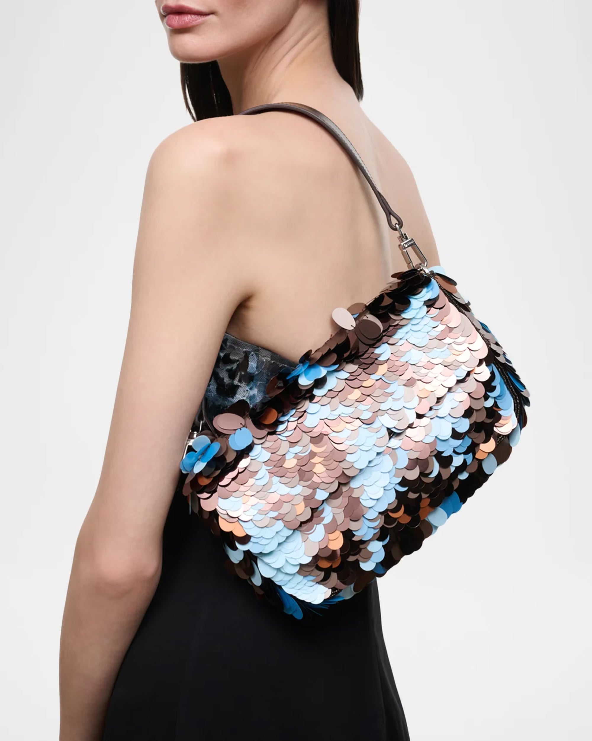 A woman wearing a strapless black dress carries a large shoulder bag covered in overlapping metallic sequins in silver, rose gold, and light blue, against a plain light gray background.