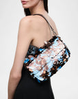 A woman wearing a strapless black dress carries a large shoulder bag covered in overlapping metallic sequins in silver, rose gold, and light blue, against a plain light gray background.