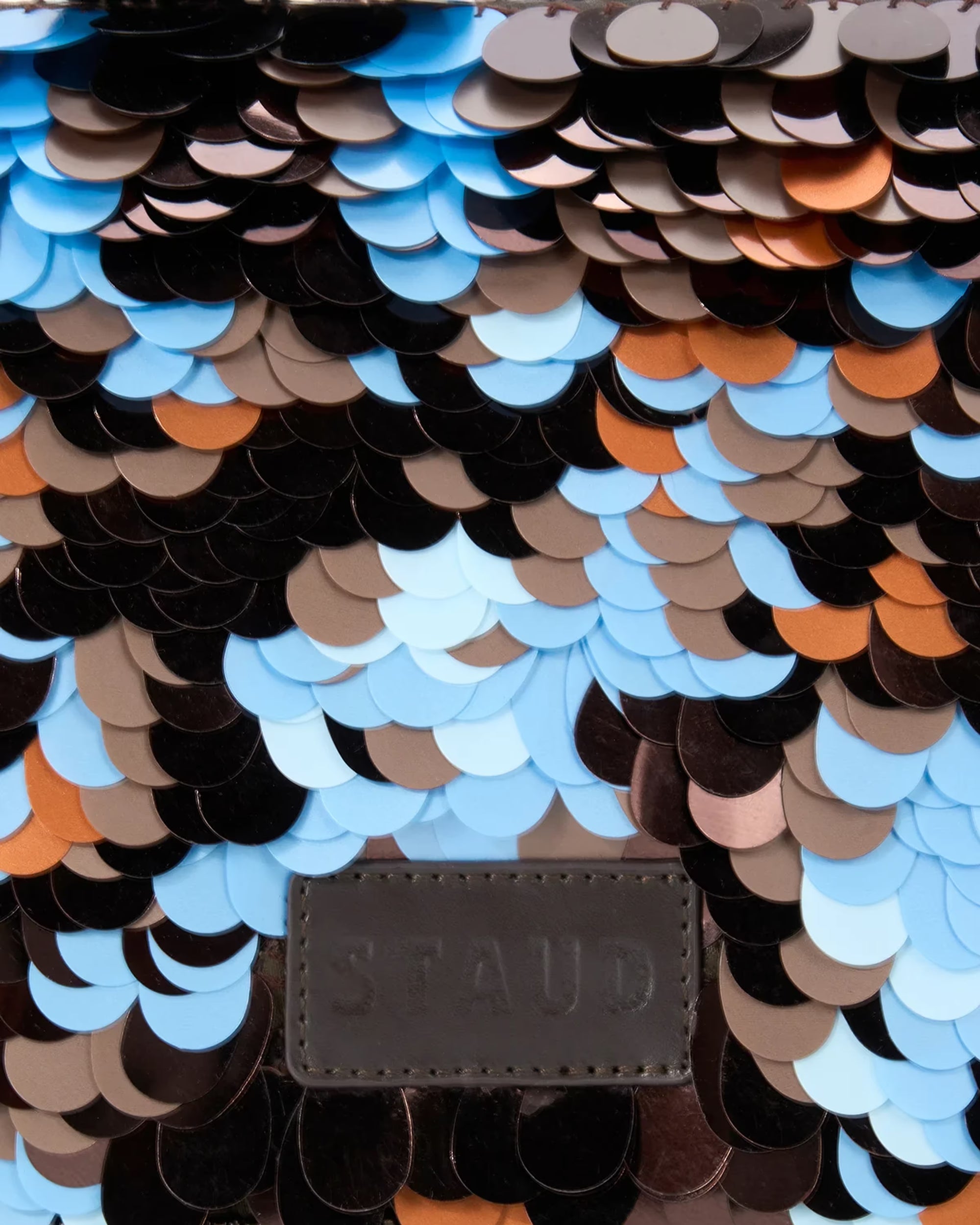 Close-up of a sequined surface with overlapping round scales in black, brown, tan, light blue, and orange, with a dark brown embossed STAUD leather patch near the bottom center.