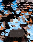 Close-up of a sequined surface with overlapping round scales in black, brown, tan, light blue, and orange, with a dark brown embossed STAUD leather patch near the bottom center.