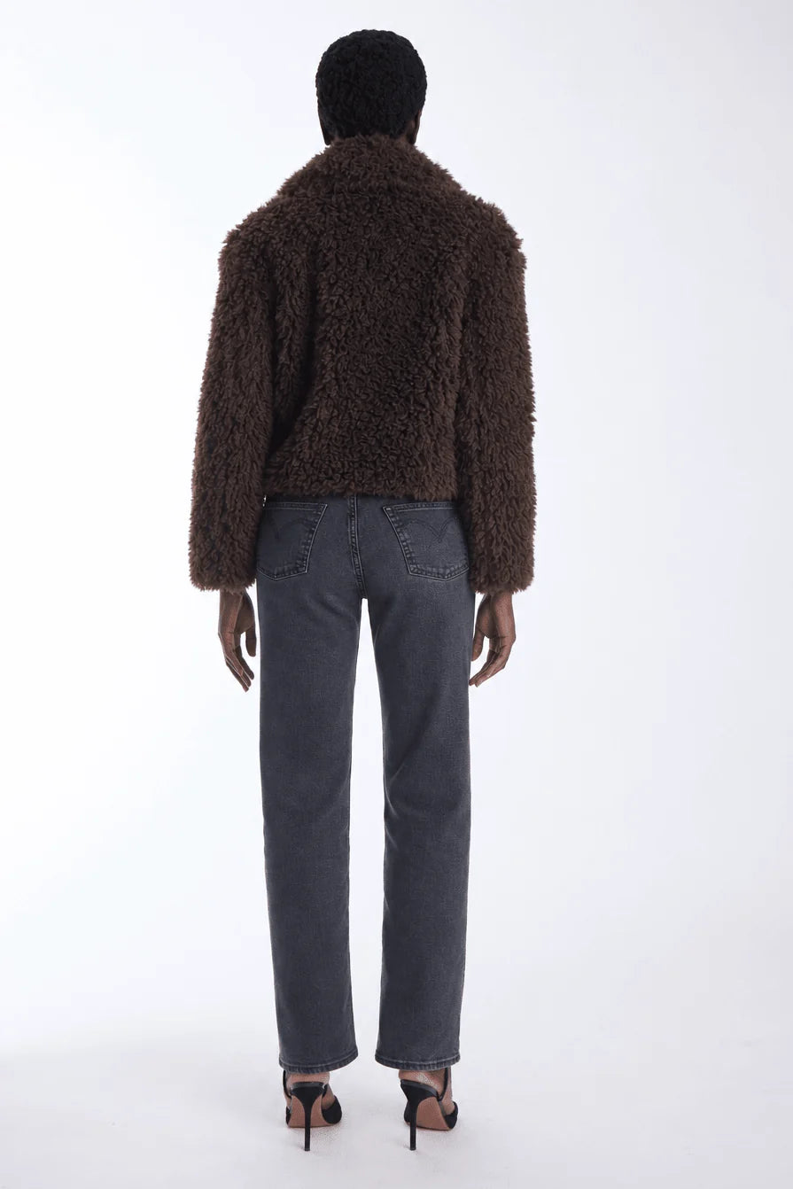 A model viewed from the back wearing a dark brown textured teddy-fur jacket, dark gray straight-leg jeans, and black high-heeled sandals against a plain white studio background.
