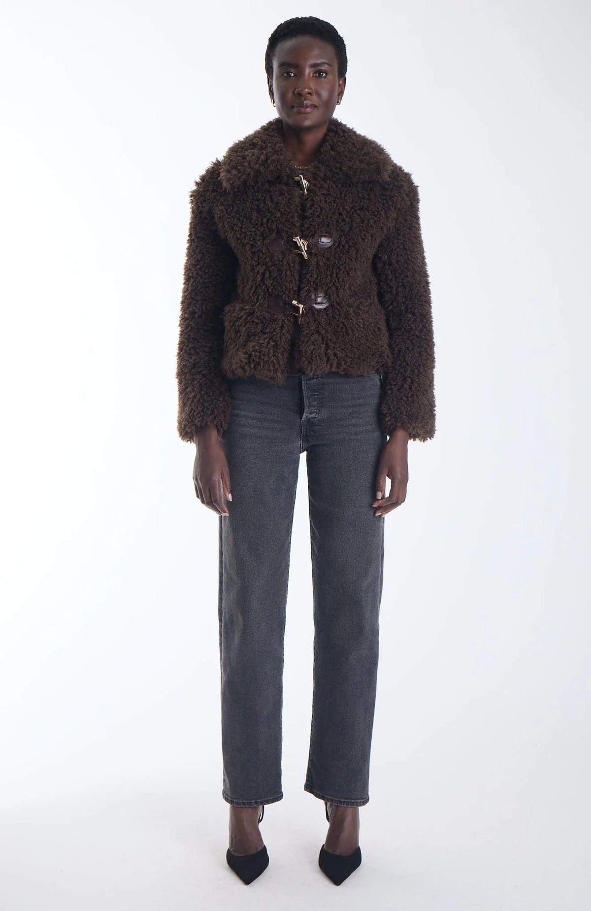 A model stands against a white studio background wearing a dark brown textured teddy-fur jacket with gold-tone buttons, dark gray straight-leg jeans, and black pointed-toe heels.