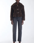 A model stands against a white studio background wearing a dark brown textured teddy-fur jacket with gold-tone buttons, dark gray straight-leg jeans, and black pointed-toe heels.