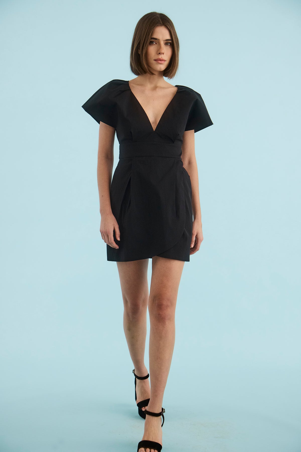 a model wearing a black short dress with black heels with a blue background 