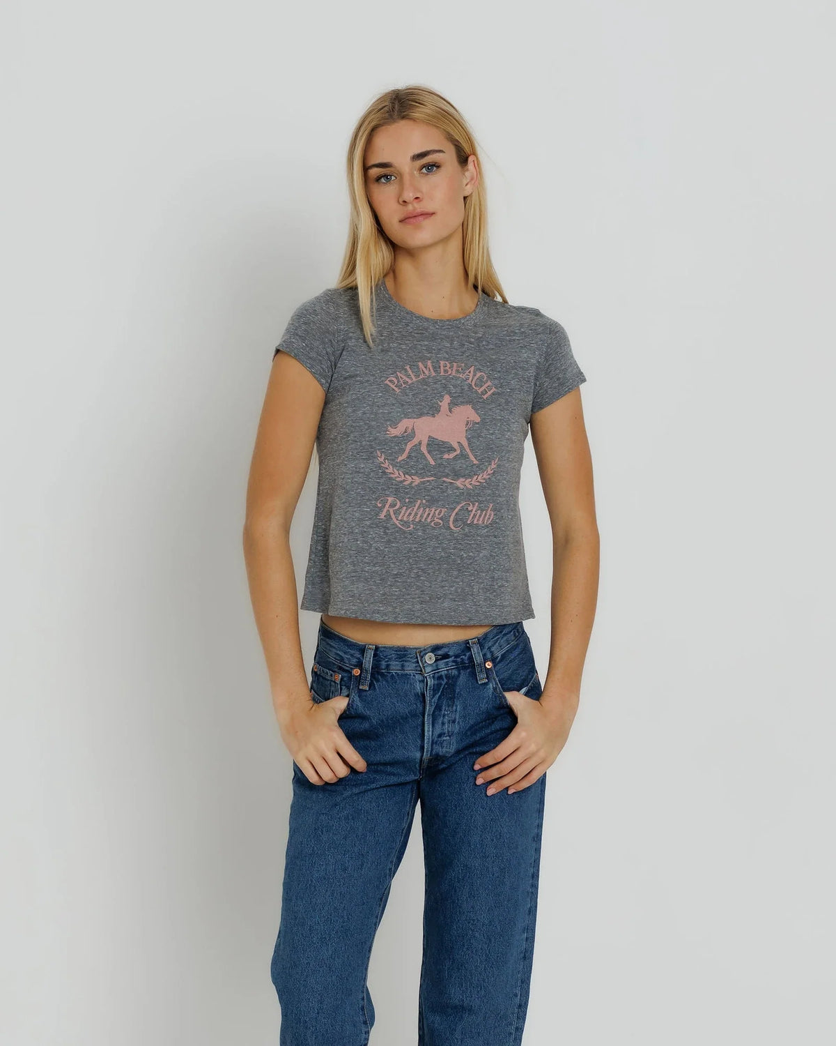 A blonde woman modeling a gray heather cropped t-shirt with a pink graphic of a horse and rider and the text 'PALM BEACH RIDING Club', paired with blue jeans, against a light gray studio background.