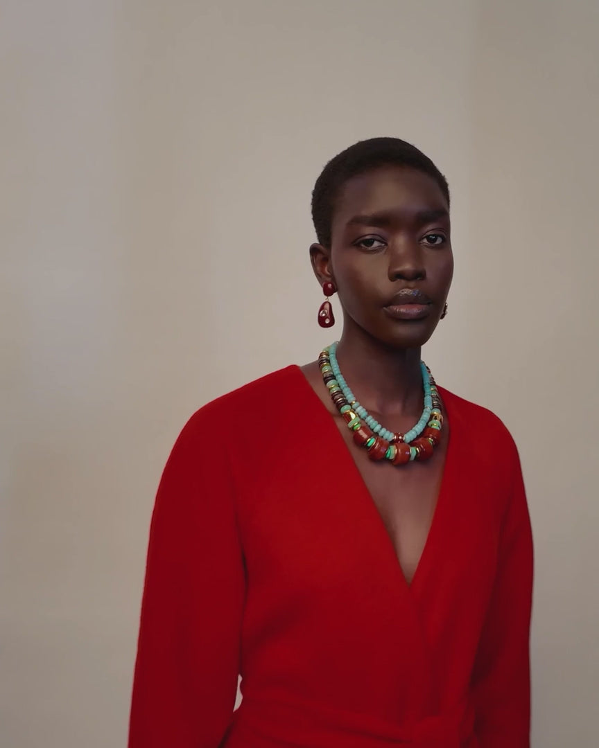 Video of a model wearing a multi-strand turquoise and brown beaded necklace with matching drop earrings, paired with a bright red top against a light beige background.