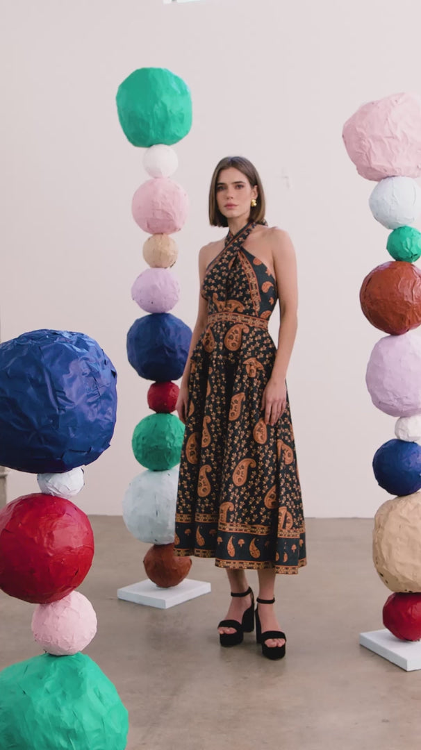 A woman models a halter-neck midi dress with a black base and brown paisley pattern, standing in a studio decorated with colorful paper lanterns.