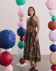 A woman models a halter-neck midi dress with a black base and brown paisley pattern, standing in a studio decorated with colorful paper lanterns.