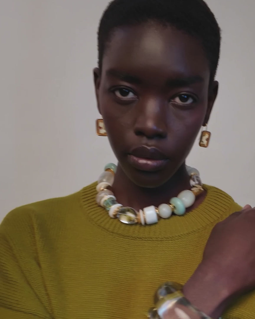 Video of a  model with short dark hair is shown from the chest up, wearing a textured olive green knit sweater, a chunky beaded necklace with white, cream, and light green beads, gold-toned earrings, and a clear acrylic bangle. The background is a clean, solid off-white studio backdrop.