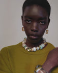 Video of a  model with short dark hair is shown from the chest up, wearing a textured olive green knit sweater, a chunky beaded necklace with white, cream, and light green beads, gold-toned earrings, and a clear acrylic bangle. The background is a clean, solid off-white studio backdrop.