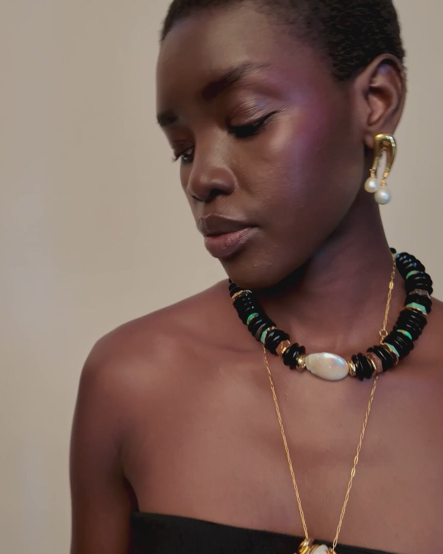 Video of a model wearing a multi-strand necklace with white pearls, black beads, and gold-toned accents, along with matching gold hoop earrings and a gold pendant necklace, styled with a black strapless top against a light beige background.