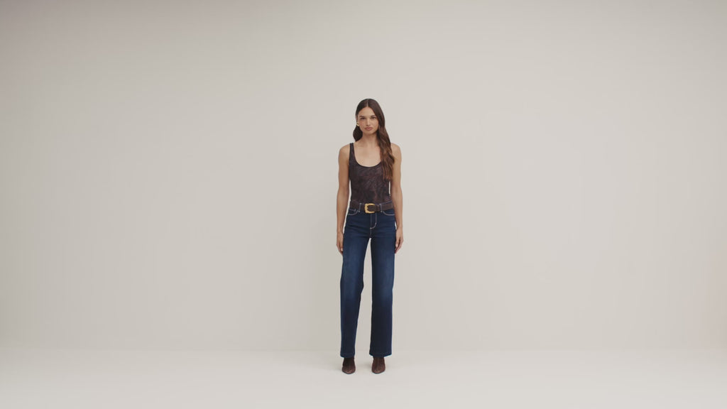 A woman stands against a plain light beige studio background wearing a sleeveless dark patterned top with a gold belt and dark blue wide-leg jeans.