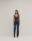 A woman stands against a plain light beige studio background wearing a sleeveless dark patterned top with a gold belt and dark blue wide-leg jeans.