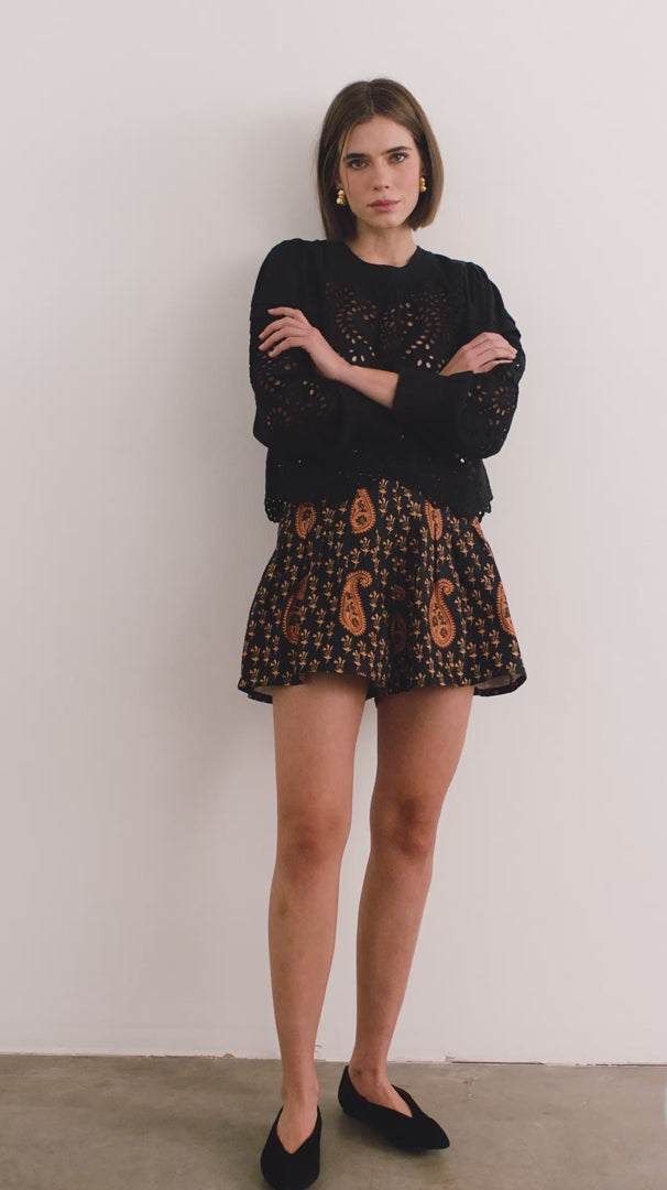 video of A woman standing in a studio wearing a black long-sleeve lace-trim top and a black shorts with a brown paisley pattern. 