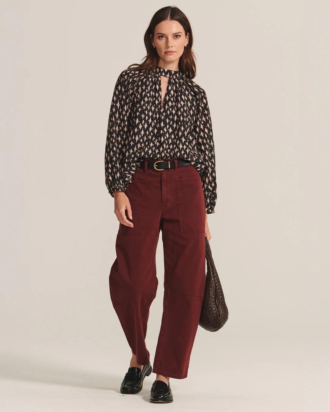 Woman wearing a patterned blouse and red pants holding a woven bag on a plain background