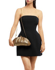 A woman modeling a strapless black mini dress and holding a metallic gold pleated clutch bag with a gold chain strap against a plain white background.