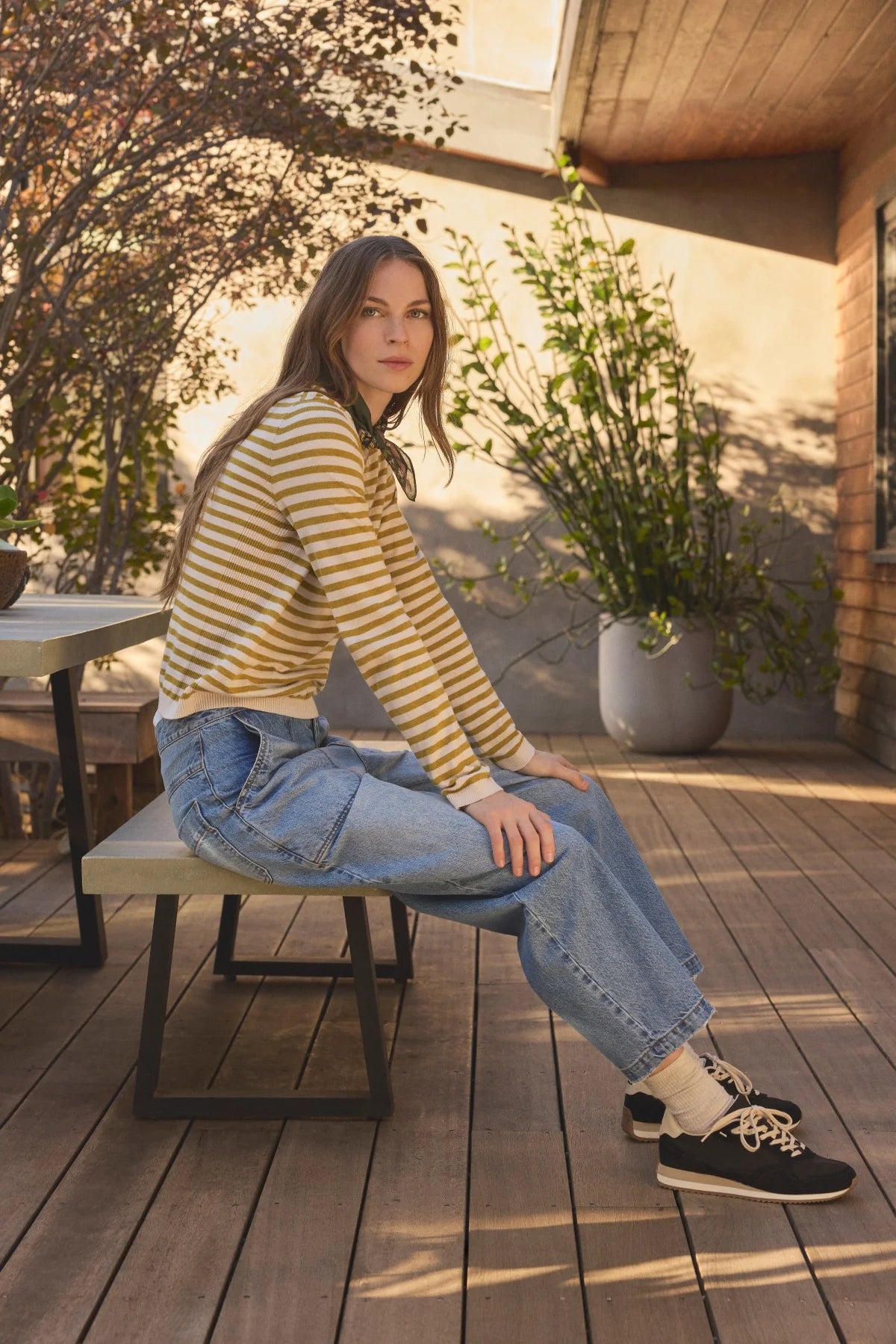 A woman sits on a wooden bench on a sunlit deck, wearing a yellow and beige striped long-sleeve top, light blue jeans, white socks, and black sneakers.