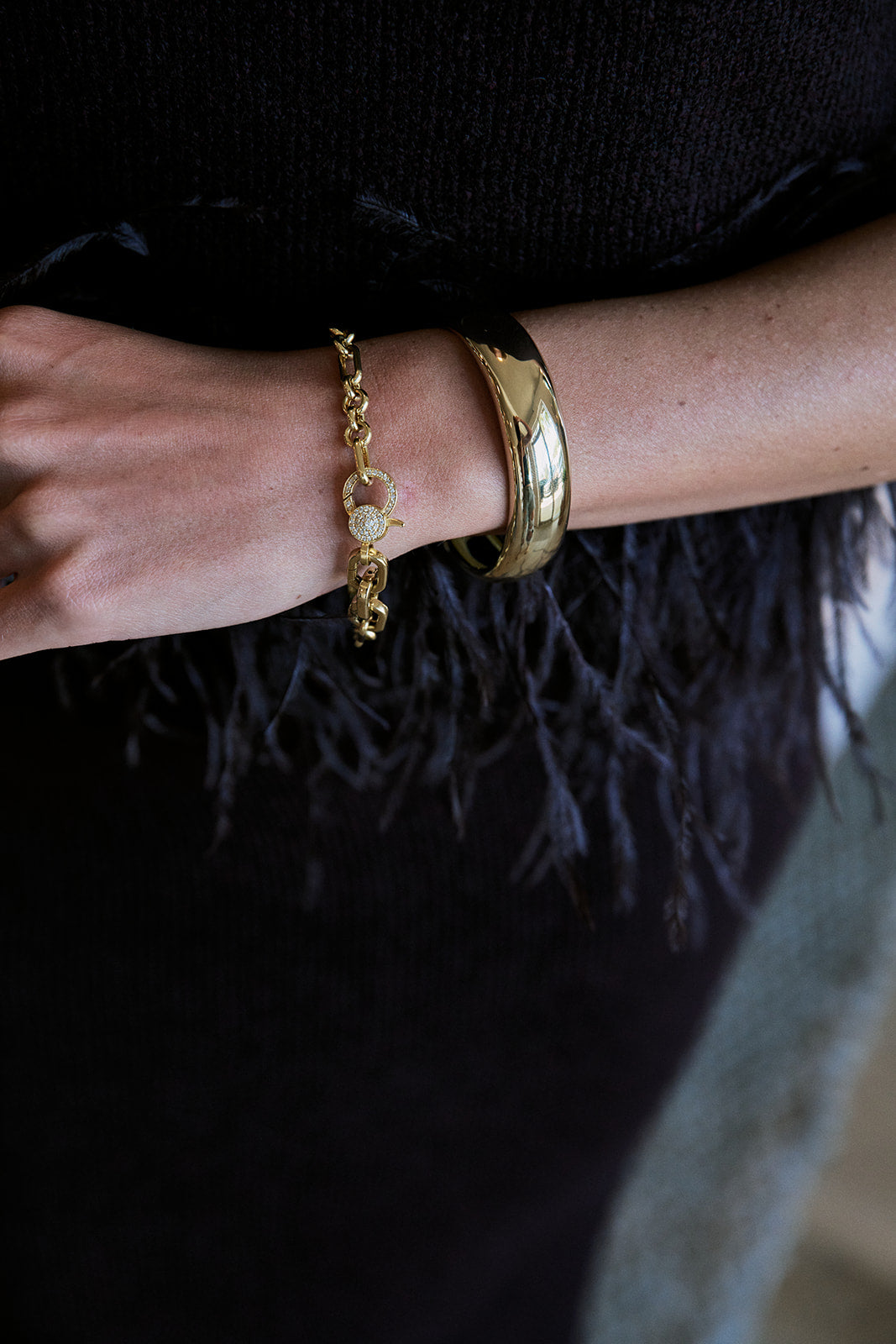 A close-up of a person's wrist wearing two gold bracelets: a delicate chain bracelet with a small round charm and a wide, polished gold bangle.
